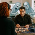 A client sits at a tarot reading table across from a medium during spiritual coaching, with two humorous ghost figures peering over the client’s shoulders at the tarot cards—symbolizing intuitive coaching with spirit support.